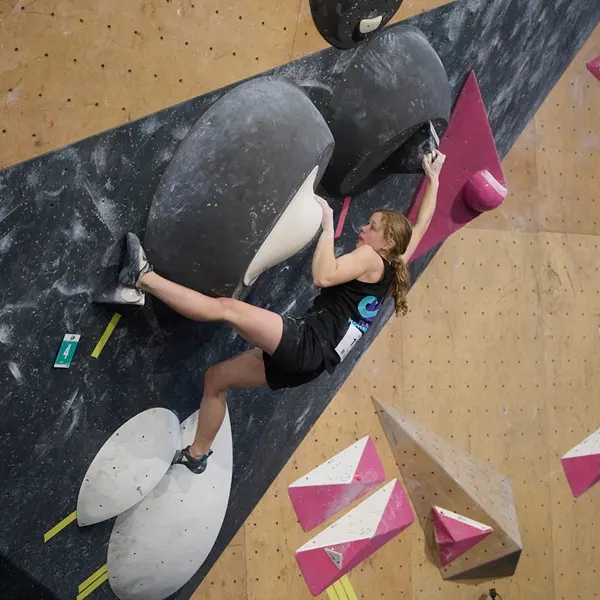 Een klimmer met lang haar, gekleed in een zwart hemdje en een korte broek, grijpt grote zwart-witte grepen vast op een indoor-klimwand; ze reikt met één arm omhoog terwijl haar benen tegen de wand drukken om haar evenwicht te bewaren.